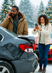 After an accident, a man speaks on the phone while a woman takes photos of the damage with her cell phone. They are near cars on a snowy day with Christmas trees nearby.